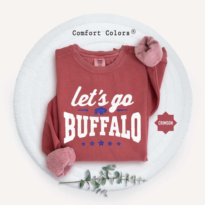 May include: A crimson long-sleeve sweatshirt with a crew neckline. The front features white text that reads let's go BUFFALO, a small blue buffalo graphic, and five blue stars. The Comfort Colors brand label is visible at the inner neck.