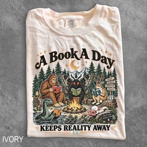 May include: An ivory t-shirt featuring a graphic print of a Bigfoot, a mothman, a jackalope, and an axolotl reading books around a campfire in a forest. The text reads A BOOK A DAY KEEPS REALITY AWAY, with a signpost saying GOOD STORIES BRIGHTER NIGHTS.
