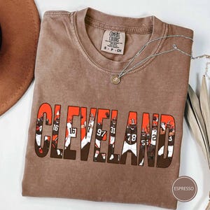 May include: A brown short-sleeve t-shirt with a crew neckline and a Comfort Colors brand label. The front features the word CLEVELAND in large orange letters, with each letter containing a graphic of a football player in a white uniform.
