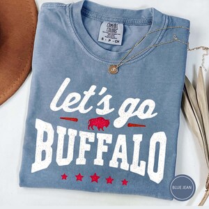May include: A blue jean colored short-sleeve t-shirt with a crew neckline. The shirt features white distressed text that reads let's go BUFFALO, a small red glitter buffalo graphic, and five red glitter stars printed across the bottom front.
