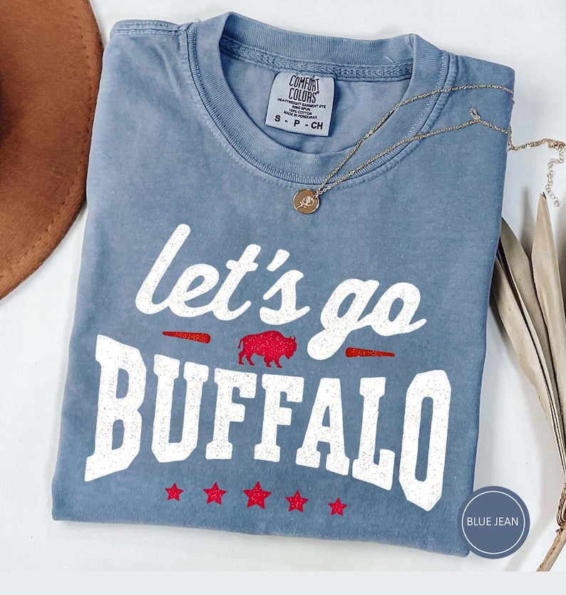 May include: A blue jean colored short-sleeve t-shirt with a crew neckline. The shirt features white distressed text that reads let's go BUFFALO, a small red glitter buffalo graphic, and five red glitter stars printed across the bottom front.