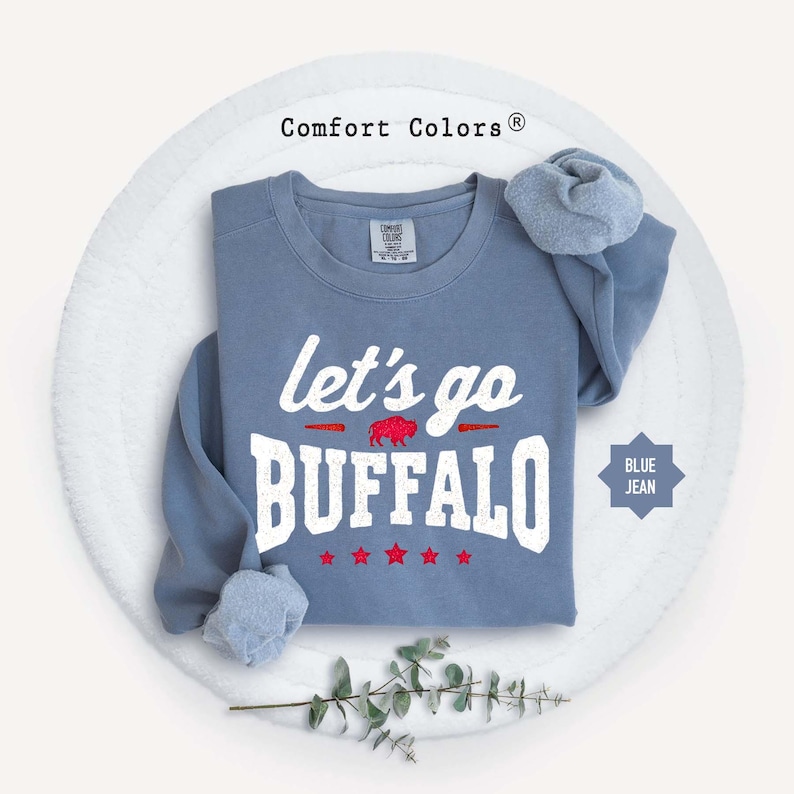 May include: A blue jean colored crewneck sweatshirt featuring the white text let's go BUFFALO with a small red buffalo graphic in the center and five red stars below. The garment has a Comfort Colors brand label at the inner neckline.