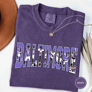 May include: A purple short-sleeved t-shirt featuring the word BALTIMORE in large block letters. Inside the letters are photographic images of football players in purple and white uniforms. The shirt has a Comfort Colors label in the neck.
