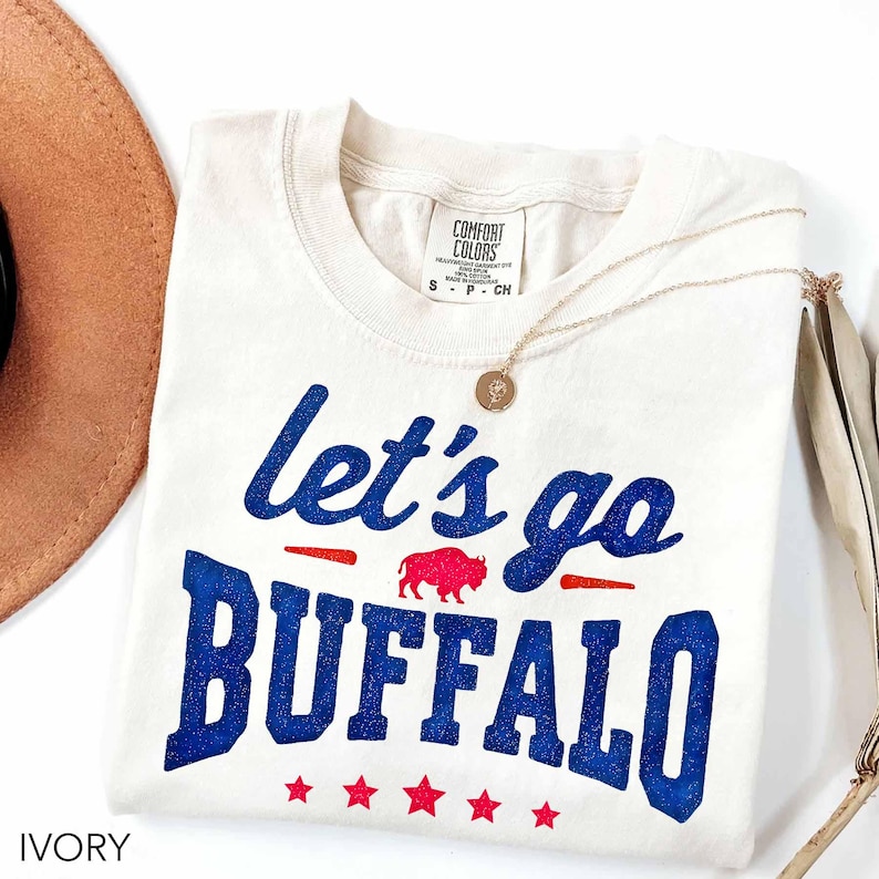 May include: An ivory short-sleeve t-shirt with a blue glitter graphic that reads "let's go BUFFALO" in a mix of script and block lettering. A small red buffalo silhouette is centered between the words, with five red stars below the text.