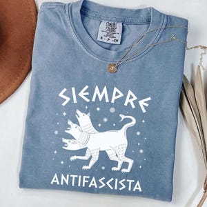 Może przedstawiać: Niebieska koszulka Comfort Colors z białymi słowami "SIEMPRE ANTIFASCISTA". Koszulka przedstawia grafikę z trójgłowym psem. Złoty naszyjnik z wisiorkiem jest zarzucony na koszulkę.
