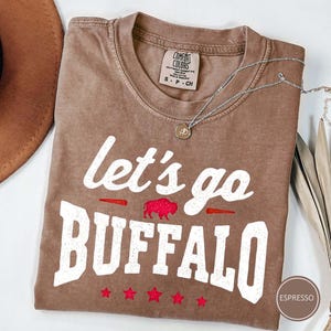 May include: An espresso-colored short-sleeve t-shirt with a crew neckline. The front features the white text "let's go BUFFALO" with a red glitter buffalo silhouette and five red glitter stars. A Comfort Colors brand label is visible at the neck.