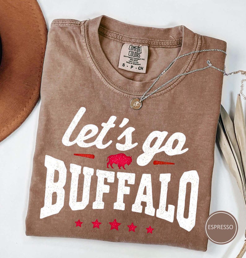 May include: An espresso-colored short-sleeve t-shirt with a crew neckline. The front features the white text "let's go BUFFALO" with a red glitter buffalo silhouette and five red glitter stars. A Comfort Colors brand label is visible at the neck.