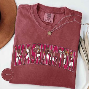 以下が含まれることがあります： チリカラーの半袖Tシャツで、大きな白いブロック体で「WASHINGTON」とプリントされています。各文字の中には、ユニフォームを着たフットボール選手のグラフィックイラストが描かれています。クルーネックで、Comfort Colorsのラベルが付いています。