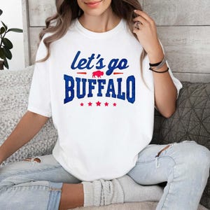 May include: A white short-sleeved t-shirt featuring the blue text let's go BUFFALO with a small red buffalo silhouette in the center. The design includes five red stars underneath the word BUFFALO, all printed on the front of the cotton shirt.