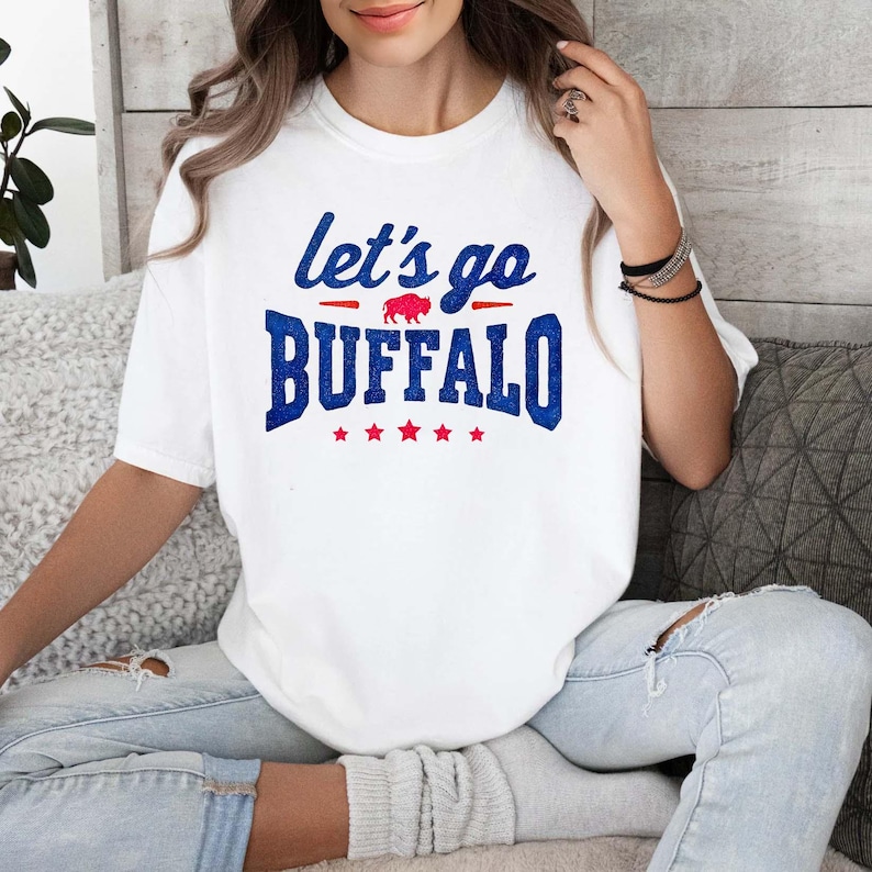 May include: A white short-sleeved t-shirt featuring the blue text let's go BUFFALO with a small red buffalo silhouette in the center. The design includes five red stars underneath the word BUFFALO, all printed on the front of the cotton shirt.