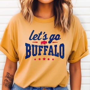 May include: A mustard yellow short-sleeve t-shirt featuring the blue glitter text "let's go BUFFALO" with a small red buffalo graphic in the center and five red stars underneath the text, all printed on the front of the garment.