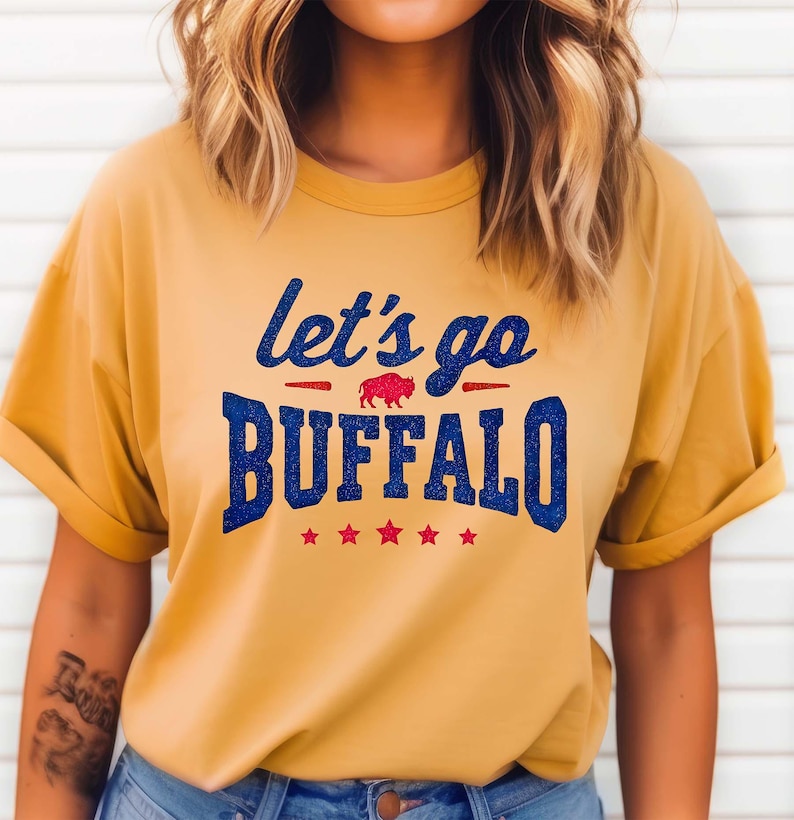 May include: A mustard yellow short-sleeve t-shirt featuring the blue glitter text "let's go BUFFALO" with a small red buffalo graphic in the center and five red stars underneath the text, all printed on the front of the garment.