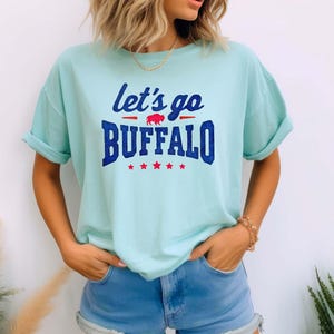 May include: A light blue t-shirt featuring the text "let's go BUFFALO" in dark blue glittery font. The design includes a small red buffalo silhouette between the words and a row of five red stars underneath the word BUFFALO on the front.