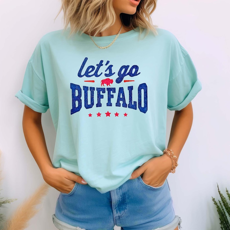 May include: A light blue t-shirt featuring the text "let's go BUFFALO" in dark blue glittery font. The design includes a small red buffalo silhouette between the words and a row of five red stars underneath the word BUFFALO on the front.