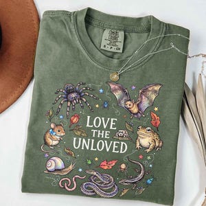 Könnte beinhalten: Ein olivgrünes T-Shirt mit einem Grafikdruck und dem Text LOVE THE UNLOVED. Das Design zeigt Illustrationen einer Vogelspinne, einer Fledermaus, einer Maus, einer Kröte, einer Schnecke, einer Schlange, einer Eidechse, eines Wurms, Spinnen, Käfern, Blättern und Sternen.