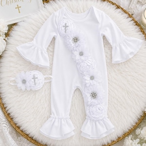 May include: A white long-sleeve romper with ruffled cuffs and a diagonal row of white fabric flowers featuring rhinestone centers and a cross charm. A matching white headband with a flower and cross charm is included with the romper.
