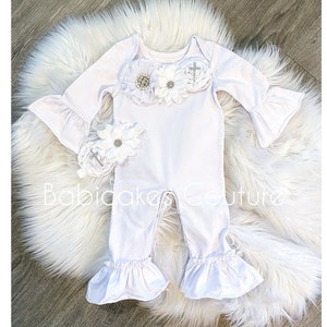 May include: White baby onesie with a ruffled hem and long sleeves. The onesie has a white flower embellishment with a silver cross in the center. The onesie is paired with a matching white flower headband.