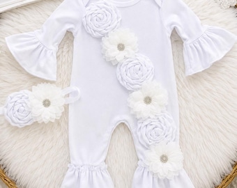 May include: A white long-sleeve romper with ruffled cuffs and a diagonal row of fabric flowers featuring rhinestone centers. A matching white headband with a fabric flower is included. A sign reads Baby's Christening with a cross symbol.