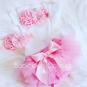 May include: Pink baby outfit with a white onesie, a pink tulle tutu, and a pink flower headband. The onesie has a pink flower applique and the text "carter's nb" on the front.