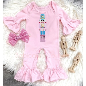 May include: A pink baby onesie with a sequined nutcracker design. The onesie has long sleeves with ruffles and a ruffled bottom. A pink sequined bow is included.
