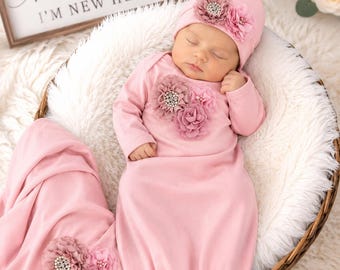 May include: A light pink long-sleeve baby gown and matching beanie set. The gown features a cluster of fabric flowers with rhinestone centers on the chest and a matching floral accent on the hat. A separate pink blanket with floral embellishments is included.