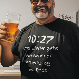 May include: A man wearing sunglasses and a black t-shirt with the text "10:27 und wieder geht ein schöner Arbeitstag zu Ende" holds a glass of beer. A clock and tools are visible in the background.