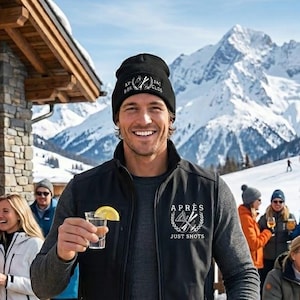 Può includere: Una persona con gilet e berretto neri tiene in mano un cocktail con una fetta di limone. Il gilet riporta la scritta "APRÈS JUST SHOTS". Lo sfondo mostra un bar di una stazione sciistica con l'insegna "APRÈS SKI CLUB" e montagne innevate.