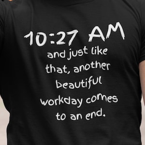 May include: Black t-shirt with the text "10:27 AM and just like that, another beautiful workday comes to an end." The person is wearing a green hat and holding a clear plastic cup with green liquid.
