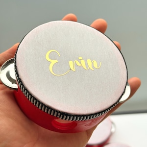 May include: A red tambourine with a white top and the name "Erin" in gold script. The instrument has a black and white striped trim and silver jingles. A musical instrument.
