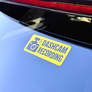 Può includere: Un adesivo rettangolare giallo con le parole "DASHCAM RECORDING" in testo blu. L'adesivo presenta anche un'icona della fotocamera blu. L'adesivo è attaccato al lunotto di un veicolo.