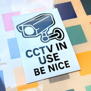 May include: A white sticker featuring a black CCTV camera illustration and the words "CCTV IN USE BE NICE". The sticker is rectangular with a shiny surface. The background has a colourful, square pattern.