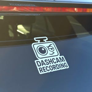 May include: White vinyl sticker with a cartoon camera design and the text "DASHCAM RECORDING". The sticker, with a winking eye, is applied to a dark blue car window. The sticker is a fun way to indicate a vehicle's dashcam.