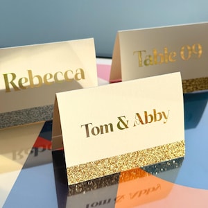 May include: Ivory place cards with gold lettering and a glittery gold or silver band. The cards display names like "Rebecca" and "Tom & Abby", and table numbers. Ideal for weddings or special events.