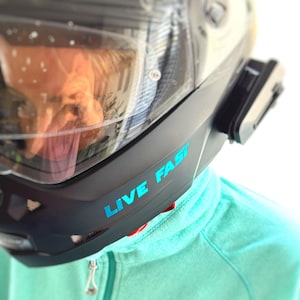 Peut inclure: Casque de moto noir avec visière transparente et l'inscription "LIVE FAST" en turquoise. Le casque est équipé d'un accessoire noir sur le côté et porté sur un sweat à capuche turquoise.