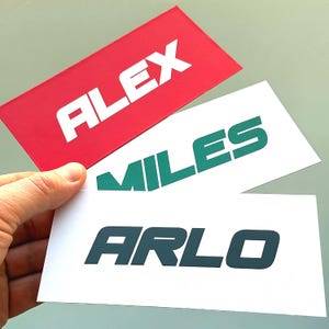 Puede incluir: Tres tarjetas rectangulares con nombres impresos. La tarjeta superior es roja con el nombre "ALEX" en blanco. La tarjeta del medio es blanca con "MILES" en verde. La tarjeta inferior es blanca con "ARLO" en verde azulado oscuro.