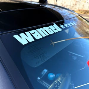 Puede incluir: Una calcomanía blanca con la palabra "Wanted" y cinco estrellas está adherida a la parte superior del parabrisas de un coche. El texto es en negrita, sin serifa, y las estrellas están alineadas a la derecha de la palabra. El interior del coche es visible.