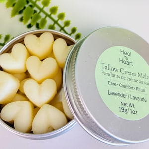 May include: A silver tin filled with heart-shaped, pale yellow tallow cream melts. The lid, slightly ajar, displays the text "Heel To Heart Tallow Cream Melts" and "Lavender/Lavande". The net weight is 19g.