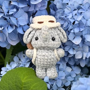 Puede incluir: Un peluche de elefante tejido a ganchillo de color gris que lleva un sombrero de pescador color crema con detalles cosidos en marrón. El elefante tiene ojos de plástico negro, orejas grandes, una trompa pequeña y una mochila color crema.