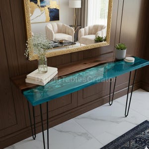 Puede incluir: Una consola moderna con una tapa de madera oscura y un diseño de río de resina epoxi turquesa. La mesa tiene patas de horquilla negras y está decorada con libros, un jarrón de flores y una pequeña planta en maceta.