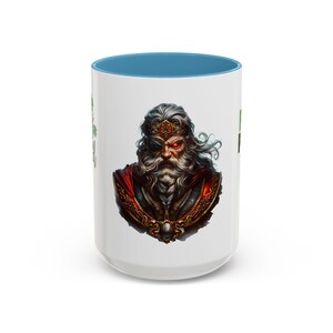May include: White ceramic mug with a blue interior. It features a detailed illustration of a bearded warrior with a red cloak, eye patch, and crown. The mug has a tall, cylindrical shape and a contrasting blue rim.