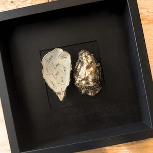 May include: A framed artwork with two oyster shells. One is gold, the other white with the word "Love" repeated in black script. The artwork is set against a black background within a black frame.