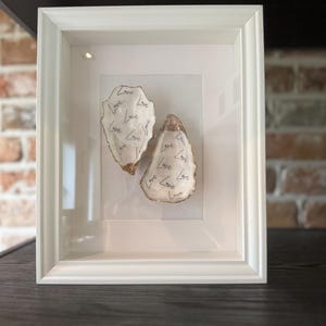 May include: A white framed artwork displaying two oyster shells. The shells are white with gold edging and the word "Love" is written repeatedly in black. The frame is white.