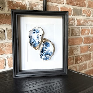 May include: A framed artwork displaying two oyster shells adorned with blue floral designs and gold edging. The shells are set against a white backdrop within a black frame, suitable for wall decor.