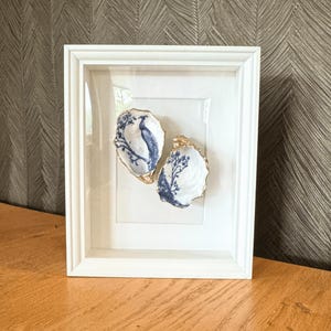 May include: A white framed artwork with two oyster shells. The shells have blue and white painted designs and gold-coloured edges. The artwork is displayed on a wooden surface. The frame is white.
