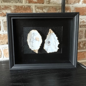May include: Framed artwork with two oyster shells, gold-edged and inscribed with text. The shells are set on a black background within a black frame. The text on the shells reads "The world is your oyster."