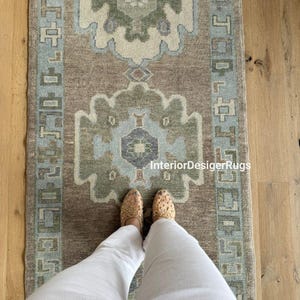 May include: A rectangular rug with a central design in shades of brown, green, and blue. The rug features geometric patterns and a border with a repeating design. The text "InteriorDesignerRugs" is visible on the rug.