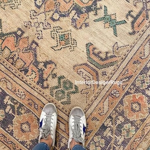 May include: A vintage-style rug with a beige background and intricate geometric patterns in shades of orange, navy, and green. The rug has a distressed look, with visible wear and tear. The image also shows a person wearing white sneakers with blue star accents.