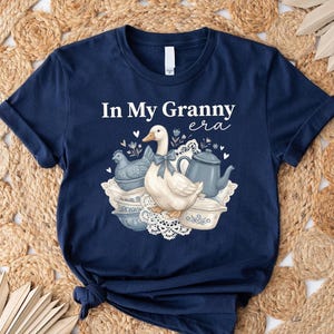An illustration of a white duck wearing a blue ribbon, surrounded by a blue hen figurine, vintage blue and white bowls, a blue kettle, and lace doilies with the text "In My Granny Era" at the top.