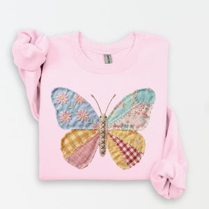 May include: A light pink sweatshirt featuring a large, patchwork butterfly design. The butterfly's wings are composed of various patterned fabrics in shades of blue, pink, yellow, and red. The butterfly has brown antennae and a stitched body.