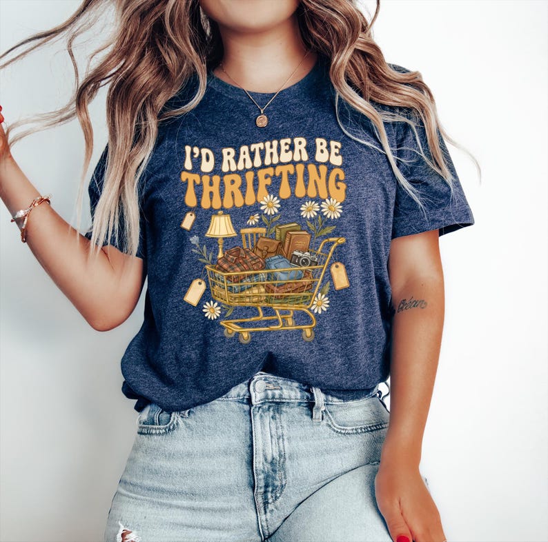 Op de afbeelding: Marineblauwe t-shirt met de tekst "I'd Rather Be Thrifting" in oranje en geel. De afbeelding toont een winkelwagentje vol vintage items, een lamp en labels, omringd door madeliefjes.